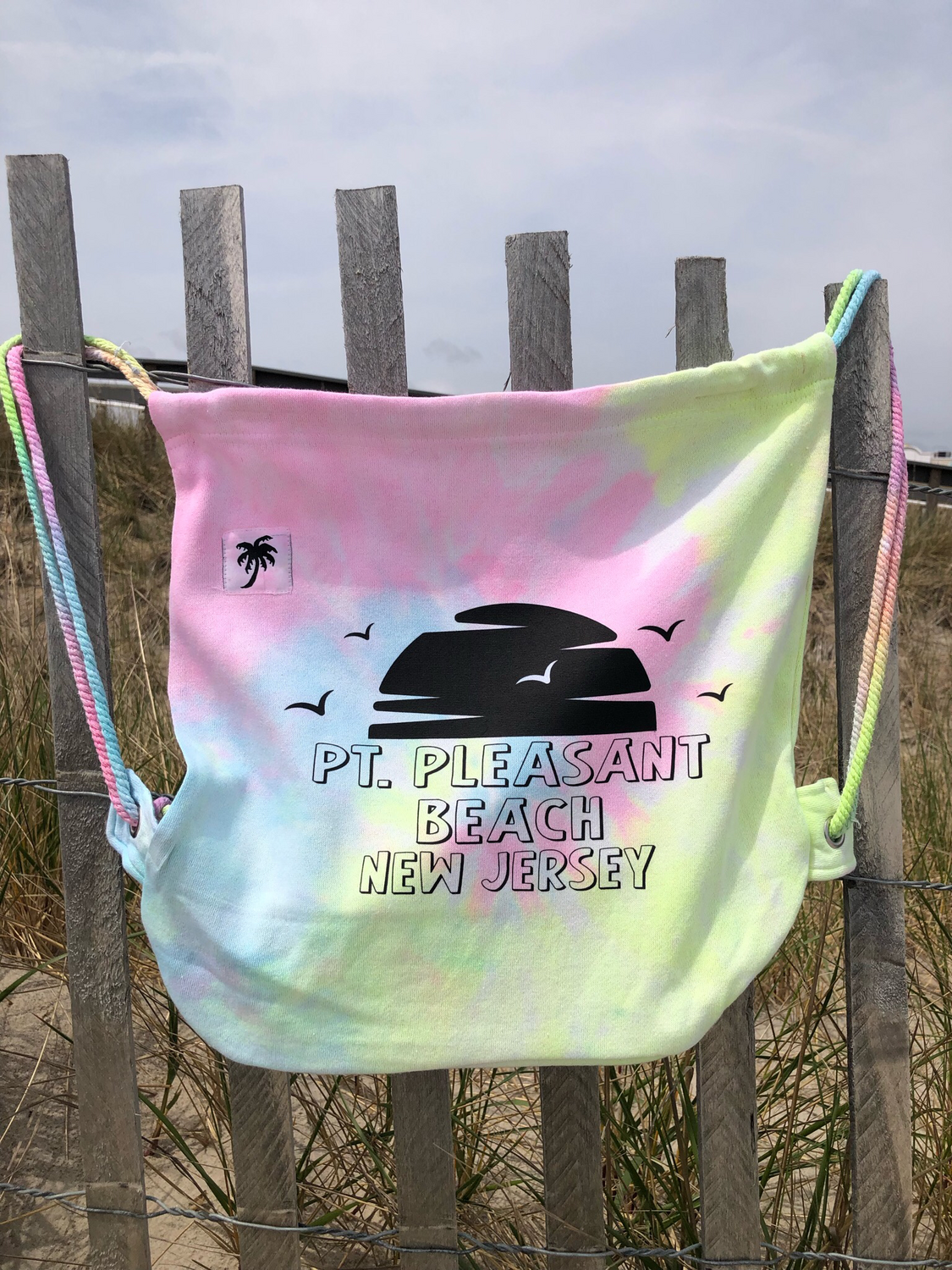 A pastel tie-dye drawstring backpack with &quot;Pt. Pleasant Beach, New Jersey&quot; text and a black sunset silhouette, displayed hanging on a wooden fence at the beach.
