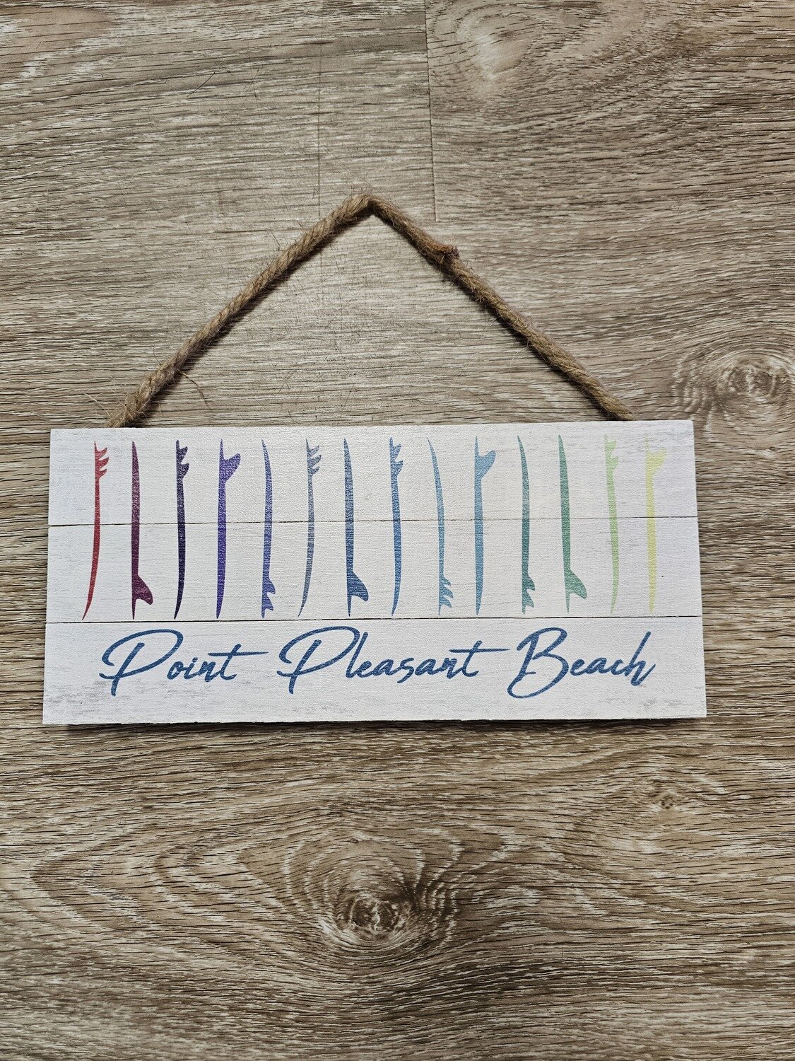 A rectangular wood sign with a drawings of surfboards lined up vertically and organized in a rainbow of colors with the words Point Pleasant Beach on the bottom