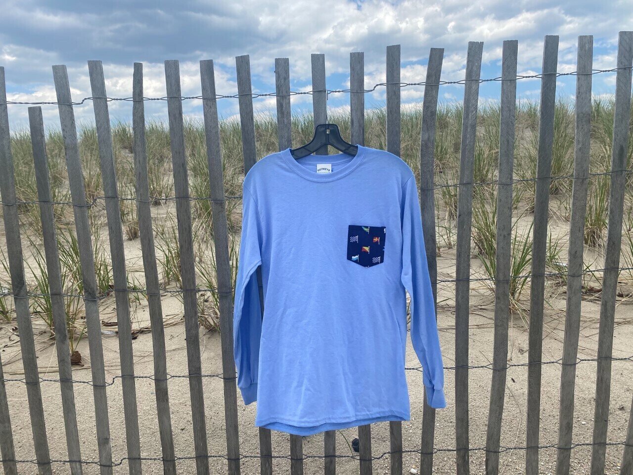 A blue long-sleeve shirt with Point Pleasant Beach detail on one sleeve and a navy pocket featuring nautical flags hanging on a wooden fence with a sandy beach and grassy dunes in the background under a cloudy sky.