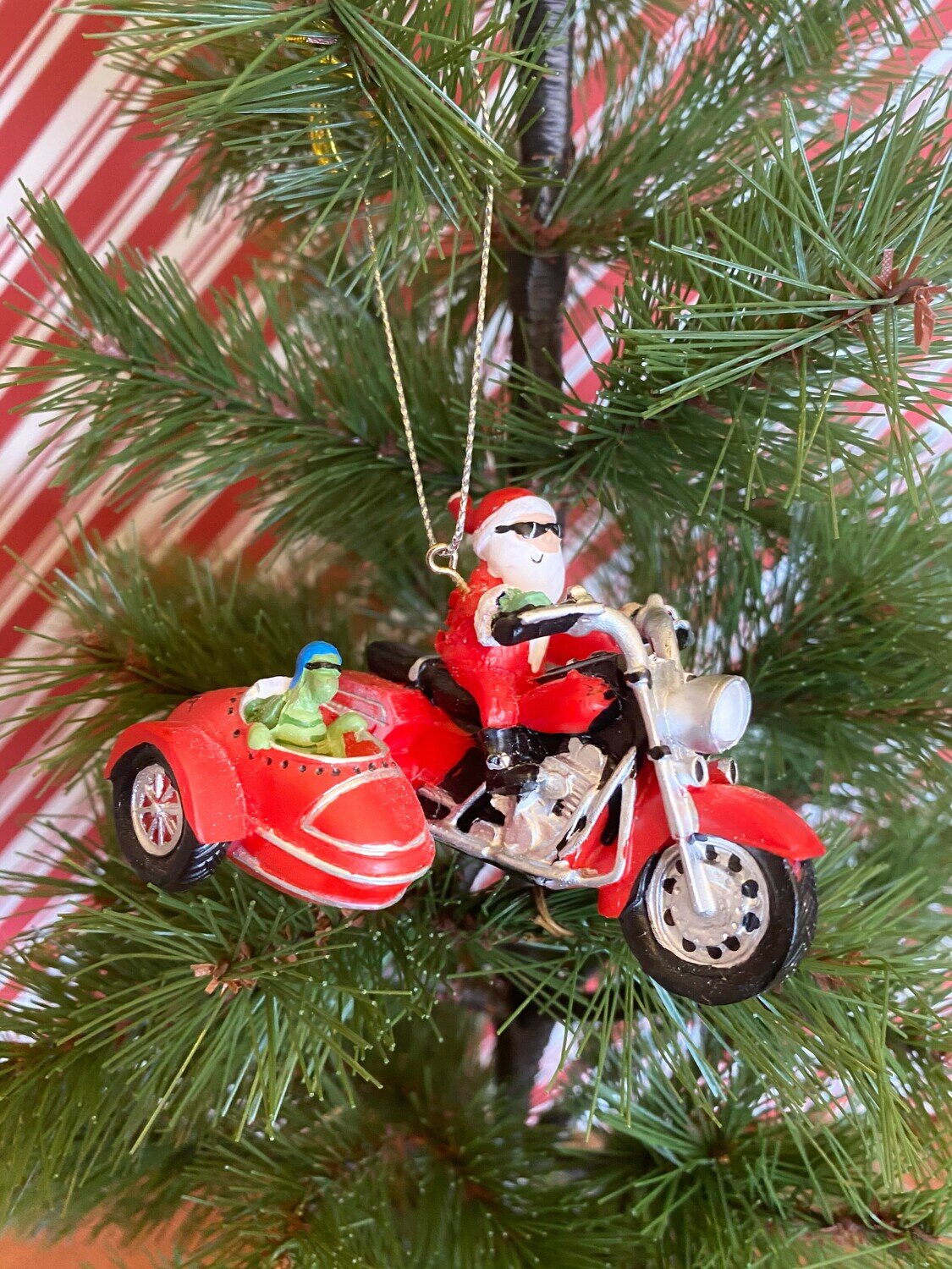 A Christmas tree ornament of Santa riding a red motorcycle with a sidecar carrying a turtle passenger, hanging on a pine tree branch.