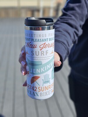 Stainless steel travel mug with a colorful design featuring text such as &quot;Greetings from Point Pleasant Beach, New Jersey,&quot; &quot;Surf,&quot; &quot;Jenkinson&#39;s,&quot; and &quot;Relax, Bike, Swim,&quot; held by a person with a boardwalk background.