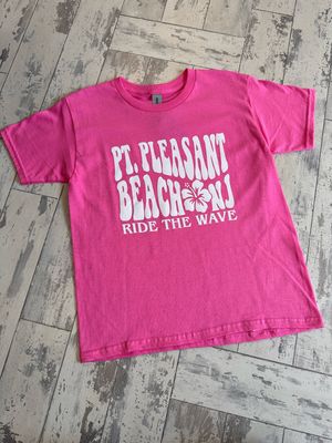 A bright pink t-shirt with &quot;Pt. Pleasant Beach NJ - Ride the Wave&quot; text and a hibiscus design in bold white font, displayed on a rustic wooden floor.