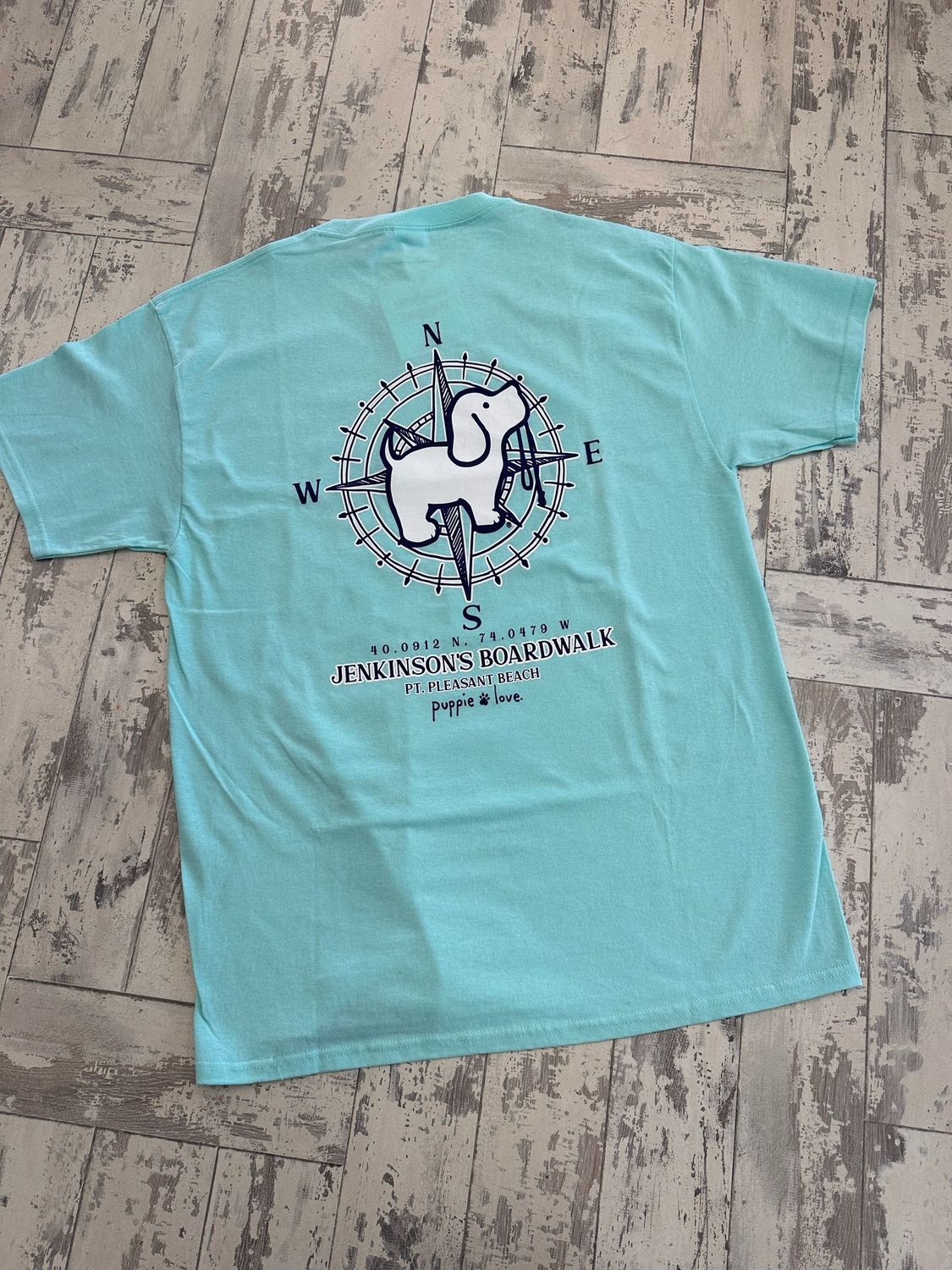 A light aqua t-shirt showcasing a navy graphic of a compass with a cartoon dog in the center, accompanied by the text &quot;Jenkinson&#39;s Boardwalk, Point Pleasant Beach, NJ&quot; below.