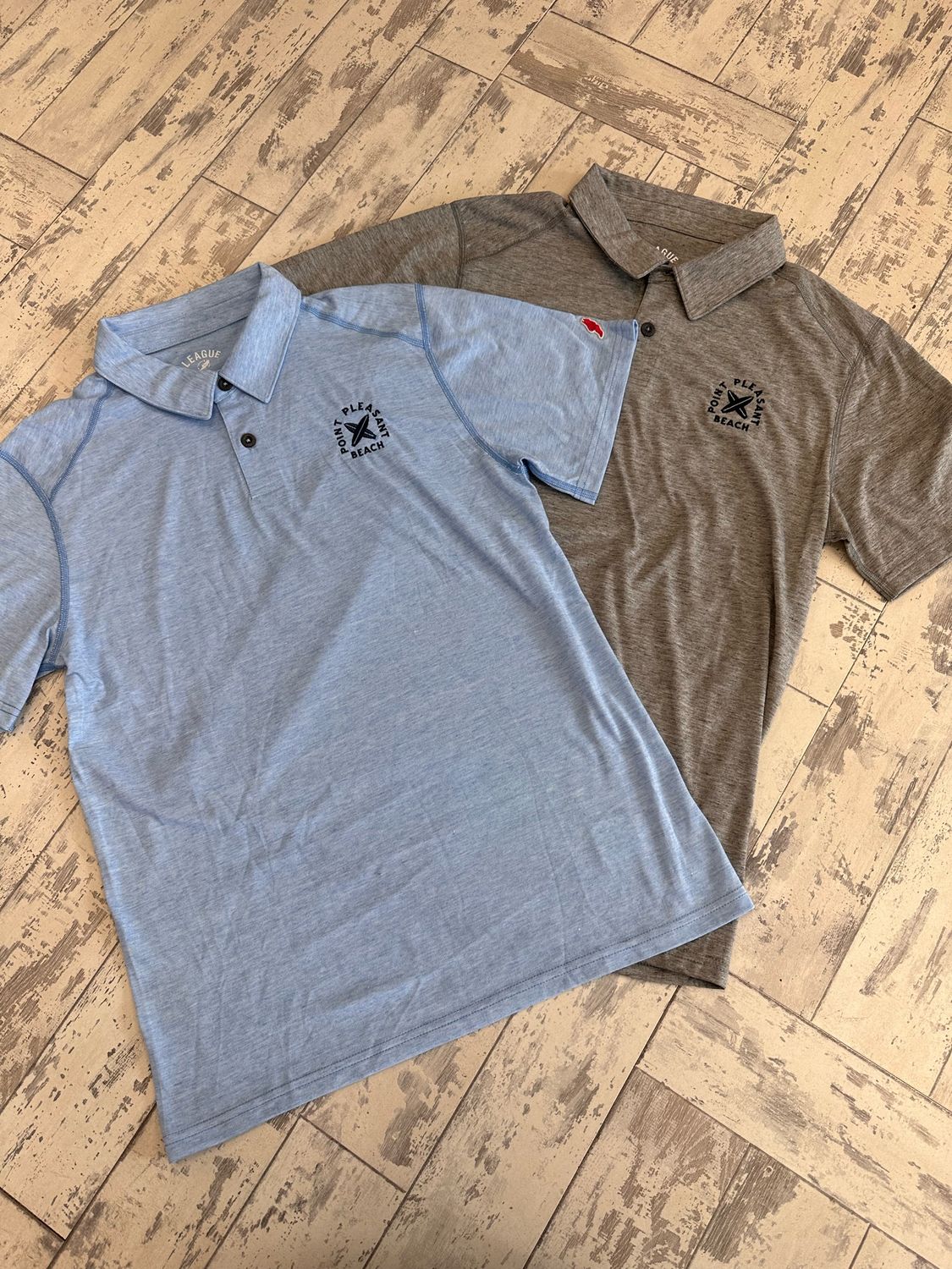 Two Point Pleasant Beach polo shirts laid flat on a wood-textured surface, one in blue and the other in graphite, featuring subtle embroidered designs on the chest.