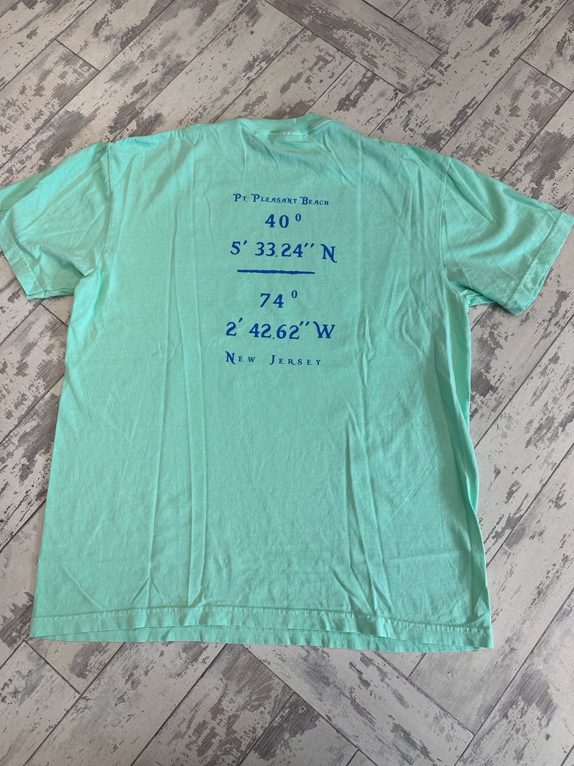 The back of a mint-green t-shirt displaying the coordinates &quot;40° 5&#39; 33.24&quot; N, 74° 2&#39; 42.62&quot; W&quot; in blue text, along with &quot;Point Pleasant Beach&quot; written above and &quot;New Jersey&quot; below.