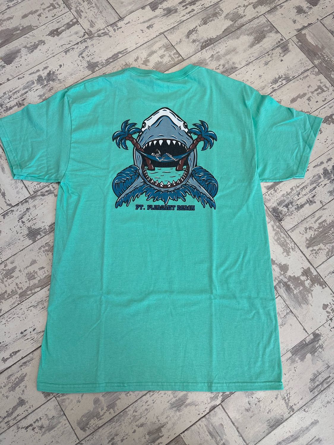 The back of the t-shirt has a skeleton drinking a bottle while sitting on a hammock as a shark appears to be ready to eat it