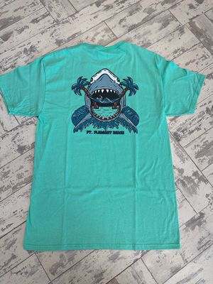 The back of the t-shirt has a skeleton drinking a bottle while sitting on a hammock as a shark appears to be ready to eat it