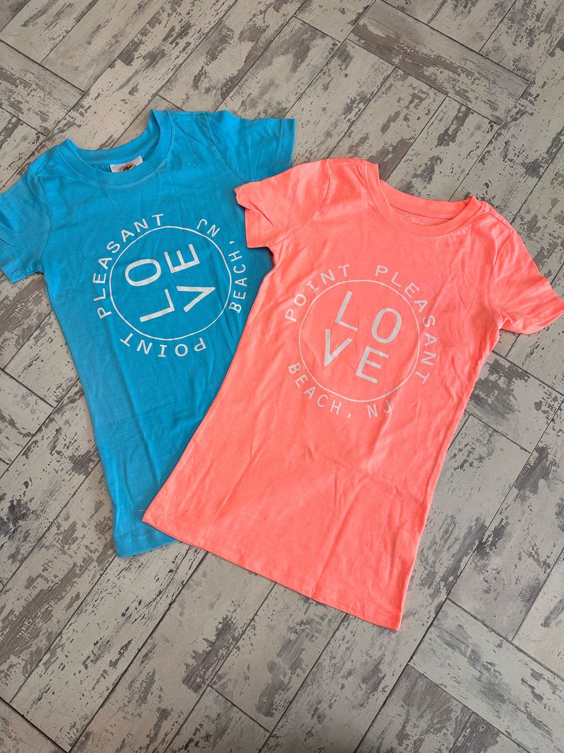 Two junior t-shirts in sky blue and coral colors with a &quot;LOVE&quot; design encircled by the text &quot;Point Pleasant Beach, NJ,&quot; displayed on a rustic wooden floor