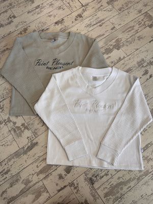Two ribbed long-sleeve shirts with &#39;Point Pleasant Beach&#39; text embroidery, one in grey and the other in white, displayed on a rustic wooden floor