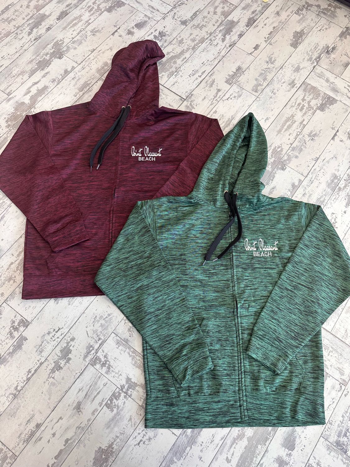 Two full-zip hooded sweatshirts with &quot;Point Pleasant Beach&quot; embroidery, one in marbled maroon and the other in marbled olive, displayed on a rustic wooden floor.