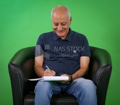 An elderly man sitting on a black chair writing notes​