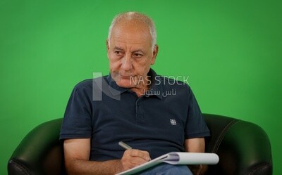 An elderly man sitting on a black chair writing notes​