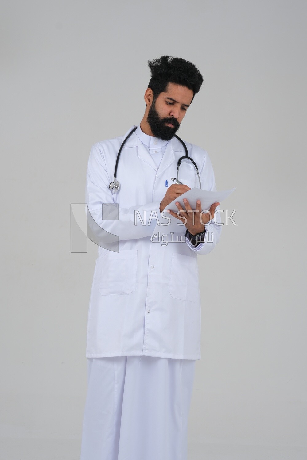 Saudi man wearing a medical coat and a stethoscope, holding a paper  to write a report on the patient&#39;s condition, white background, medicine and health care, Saudi model