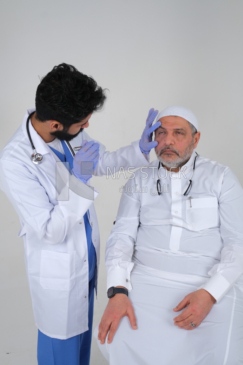 Saudi man wearing a medical coat and a stethoscope, checking the patient&#39;s eyes, white background, medicine and health care, Saudi model