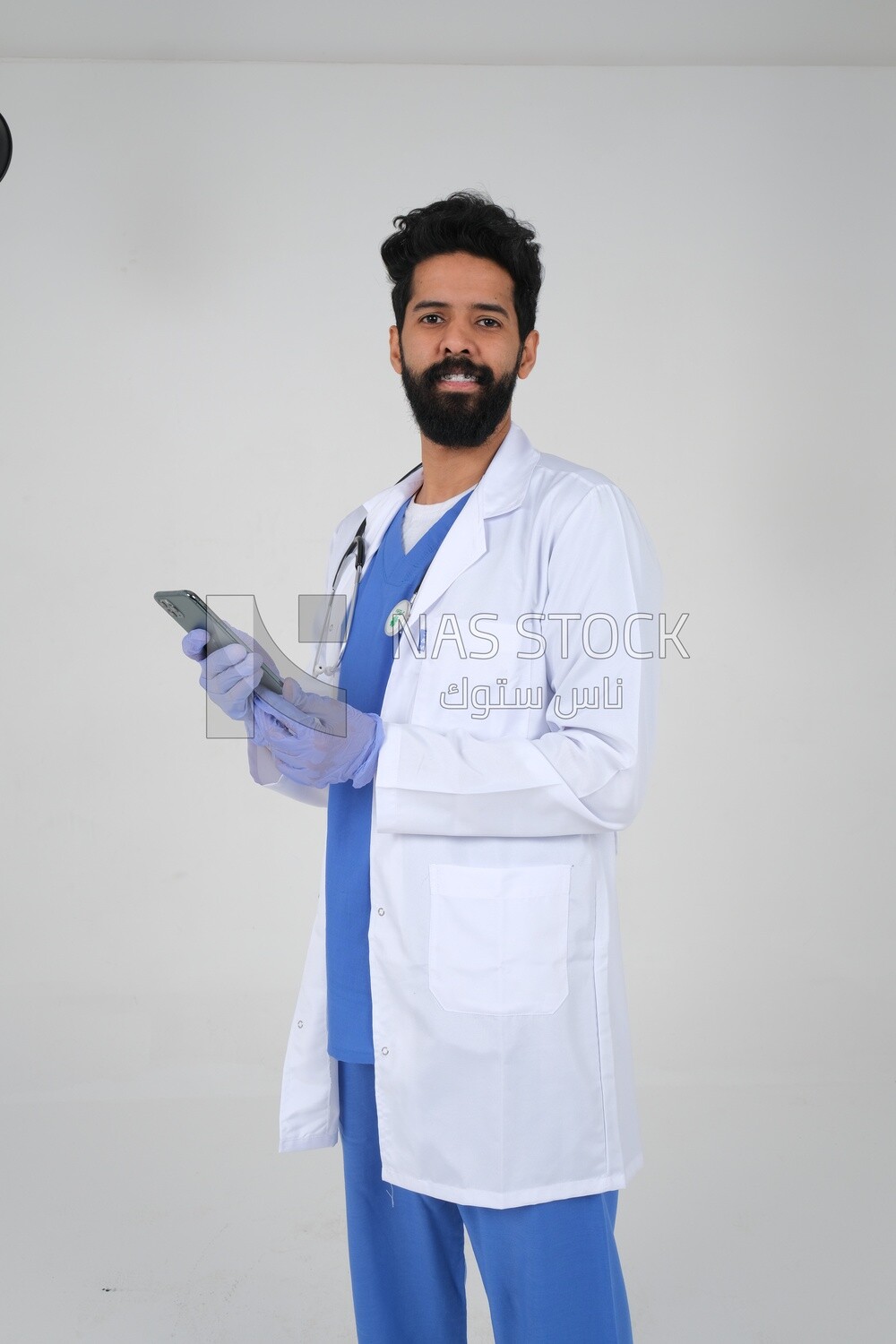 Saudi man wearing a medical coat and a stethoscope, using his mobile, white background, medicine and health care, Saudi model