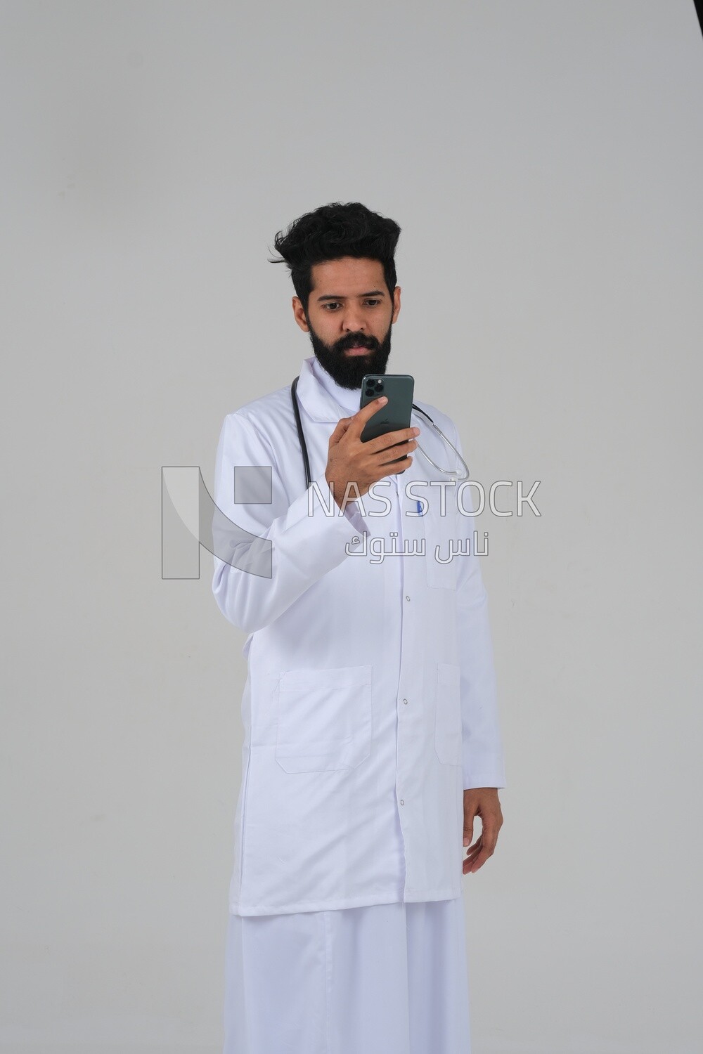 Saudi man wearing a medical coat and a stethoscope, using his mobile, white background, medicine and health care, Saudi model