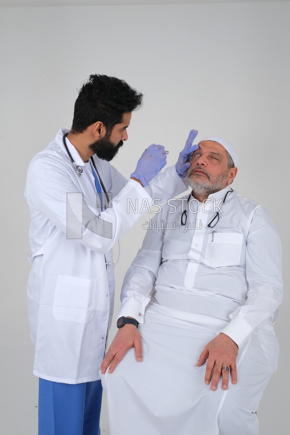 Saudi man wearing a medical coat and a stethoscope, checking the patient&#39;s eyes, white background, medicine and health care, Saudi model