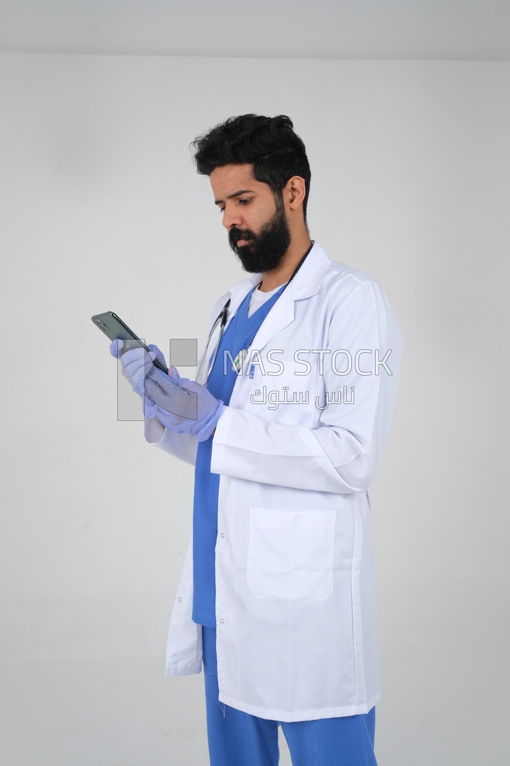Saudi man wearing a medical coat and a stethoscope, using his mobile, white background, medicine and health care, Saudi model