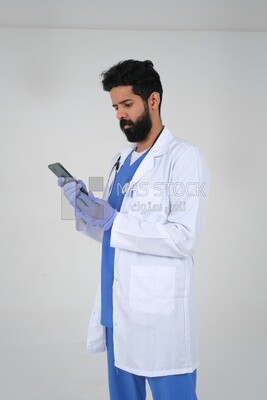 رجل سعودي يرتدي معطفًا طبيًا وسماعة طبيب يتسخدم هاتفه المحمول ، الخلفية البيضاء ، الطب والرعاية الصحية