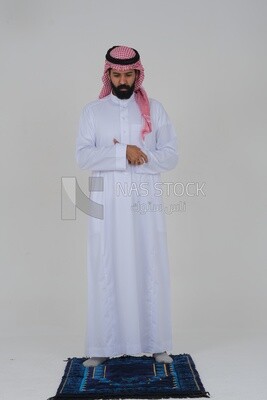Saudi man wearing a white thobe and shemagh praying on the prayer rug, praying to god, Islam and worship, white background, Saudi model