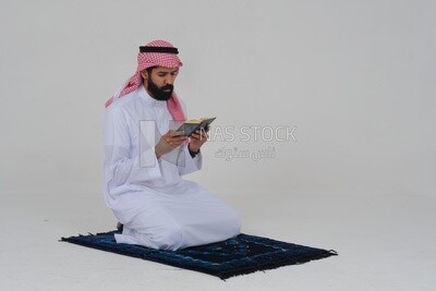 Side view of a Saudi man wearing a white dress sitting on the prayer rug, reading the Quran, praying to god, praying at the prayer rug, Islam and worship, white background, Saudi model