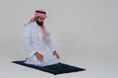 Side view of a Saudi man wearing a white dress sitting on the prayer rug, praying to god, praying on the prayer rug, Islam and worship, white background, Saudi model