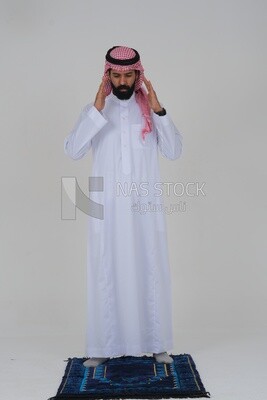Saudi man wearing a white thobe and shemagh praying on the prayer rug, praying to god, Islam and worship, white background, Saudi model