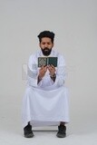 Saudi man wearing a white dress sitting on a chair, reading Quran, Islam and worship, Saudi model, white background