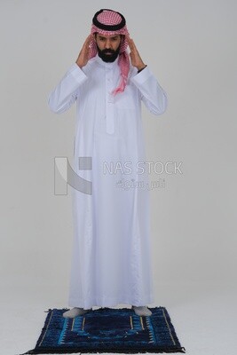Saudi man wearing a white thobe and shemagh praying on the prayer rug, praying to god, Islam and worship, white background, Saudi model
