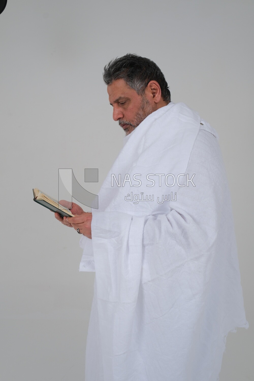 Saudi man wearing the Ihram dress and holding the Holy Quran book, praying to god, Islam and worship, performing Hajj and Umrah, remembrances and supplications, white background, Saudi model