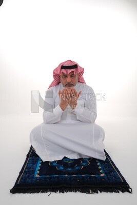 Saudi man wearing a white thobe and shemagh praying on the prayer rug, praying to god, Islam and worship, white background, Saudi model