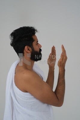 Side of a Saudi man wearing the Ihram dress and raising his hands to supplicate to God, Islam and worship, performing Hajj and Umrah, remembrances and supplications, white background, Saudi model