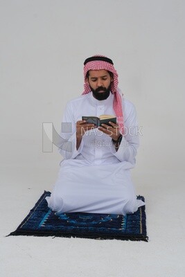 Saudi man wearing a white dress sitting on the prayer rug, reading the Quran, praying to god, praying at the prayer rug, Islam and worship, white background, Saudi model