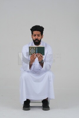 Saudi man wearing a white dress sitting on a chair, reading Quran, Islam and worship, Saudi model, white background