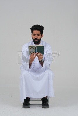 Saudi man wearing a white dress sitting on a chair, reading Quran, Islam and worship, Saudi model, white background