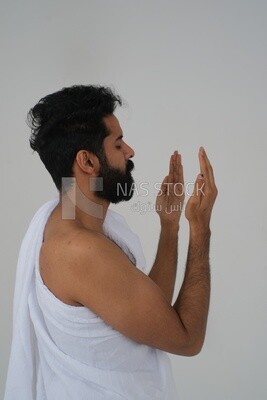 Side of a Saudi man wearing the Ihram dress and raising his hands to supplicate to God, Islam and worship, performing Hajj and Umrah, remembrances and supplications, white background, Saudi model