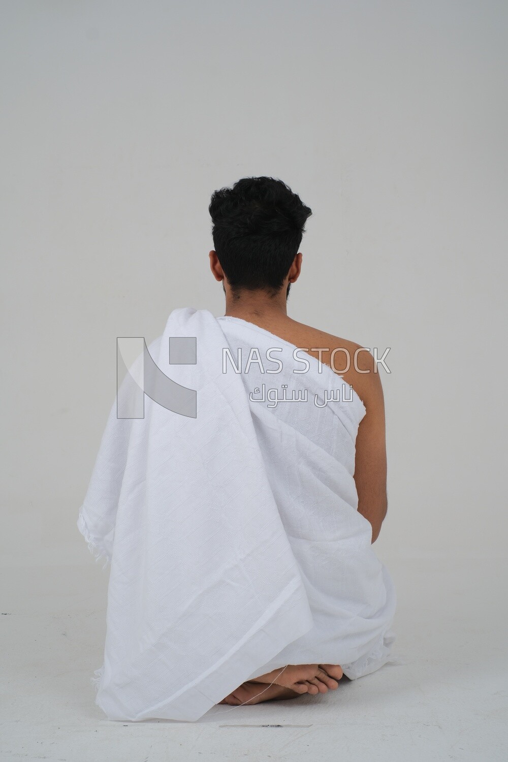 Back of a Saudi man wearing the Ihram dress sitting on the floor to supplicate to God, Islam and worship, performing Hajj and Umrah, remembrances and supplications, white background, Saudi model