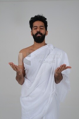 Saudi man wearing the Ihram dress and raising his hands to supplicate to God, Islam and worship, performing Hajj and Umrah, remembrances and supplications, white background, Saudi model