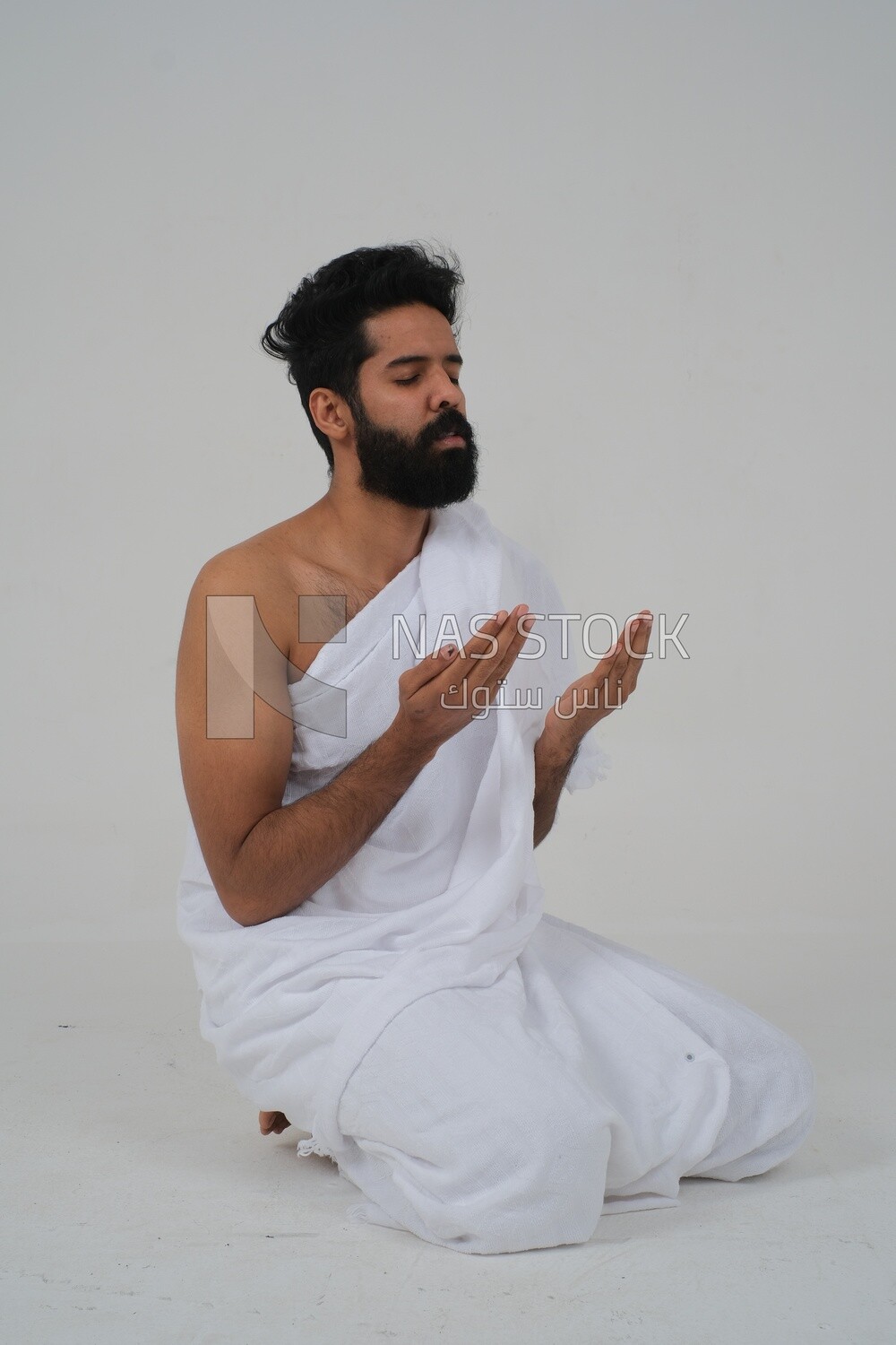 Side of a Saudi man wearing the Ihram dress and raising his hands to supplicate to God, Islam and worship, performing Hajj and Umrah, remembrances and supplications, white background, Saudi model