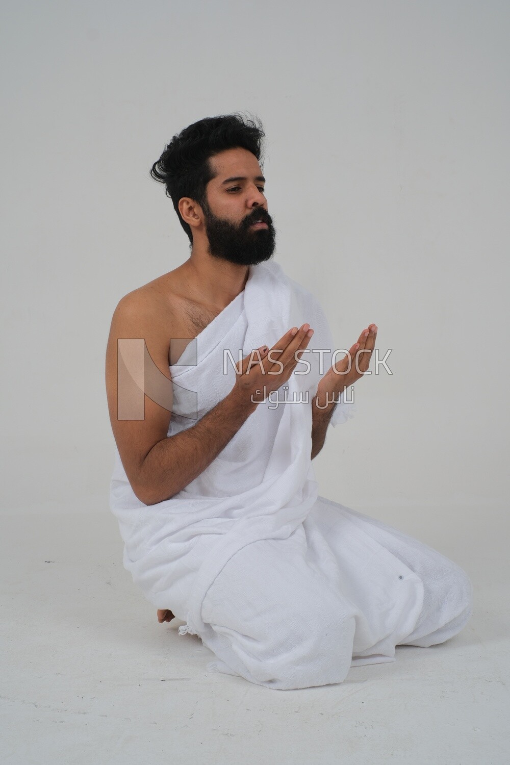 Side of a Saudi man wearing the Ihram dress and raising his hands to supplicate to God, Islam and worship, performing Hajj and Umrah, remembrances and supplications, white background, Saudi model
