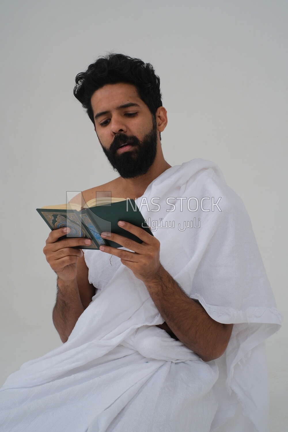 Saudi man wearing the Ihram dress and holding the Holy Quran book, praying to god, Islam and worship, performing Hajj and Umrah, remembrances and supplications, white background, Saudi model