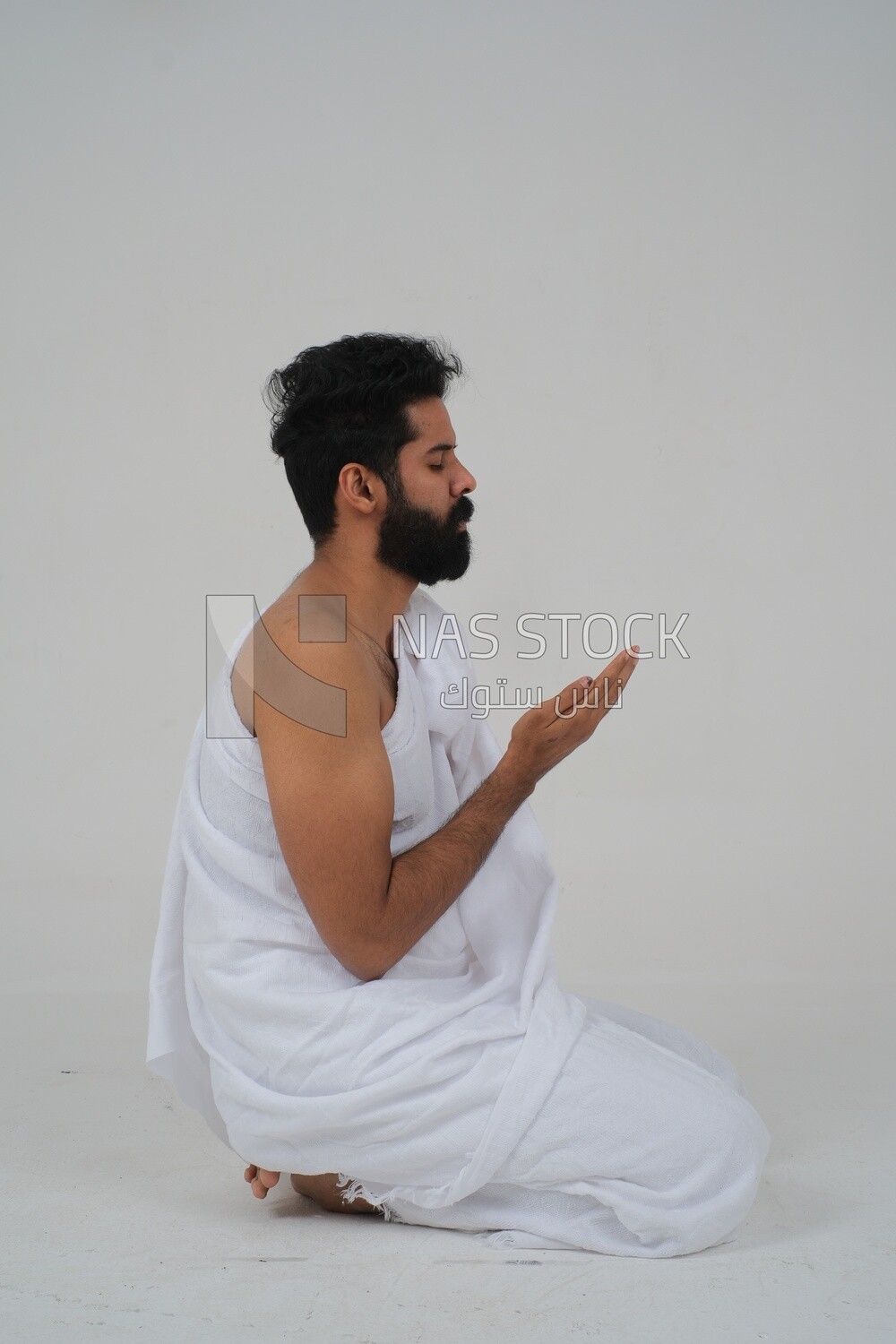 Side of a Saudi man wearing the Ihram dress and raising his hands to supplicate to God, Islam and worship, performing Hajj and Umrah, remembrances and supplications, white background, Saudi model