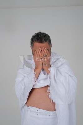 Saudi man wearing the Ihram dress puts his hand on his face to supplicate to God, Islam and worship, performing Hajj and Umrah, remembrances and supplications, white background, Saudi model