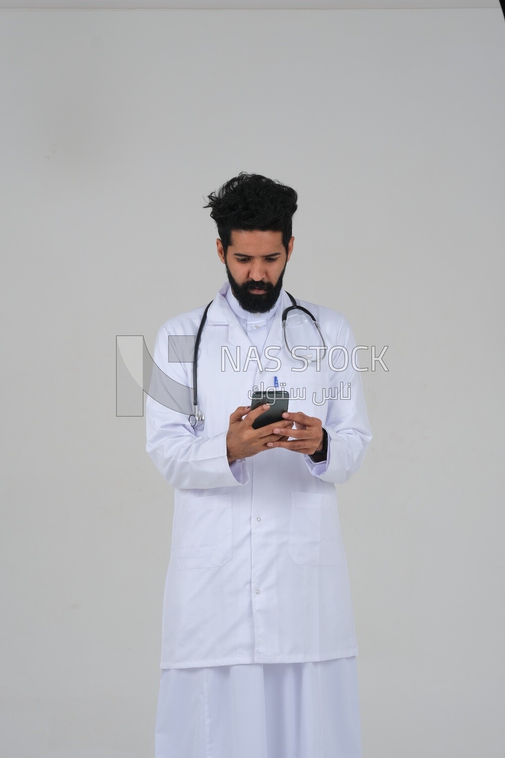Saudi man wearing a medical coat and a stethoscope, using his mobile, white background, medicine and health care, Saudi model