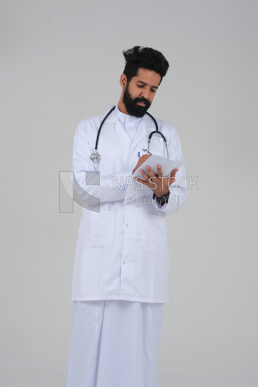 Saudi man wearing a medical coat and a stethoscope, holding a paper  to write a report on the patient&#39;s condition, white background, medicine and health care, Saudi model
