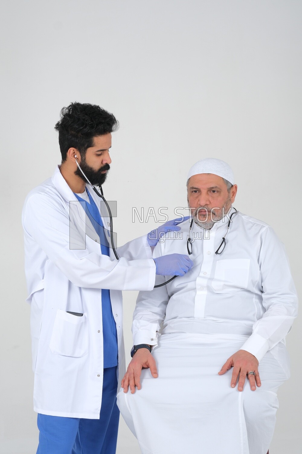 رجل سعودي يرتدي معطفًا طبيًا وسماعة طبيب ، يستمع الطبيب إلى نبضات قلب المريض ، والخلفية البيضاء ، والطب والرعاية الصحية ، موديل سعودى