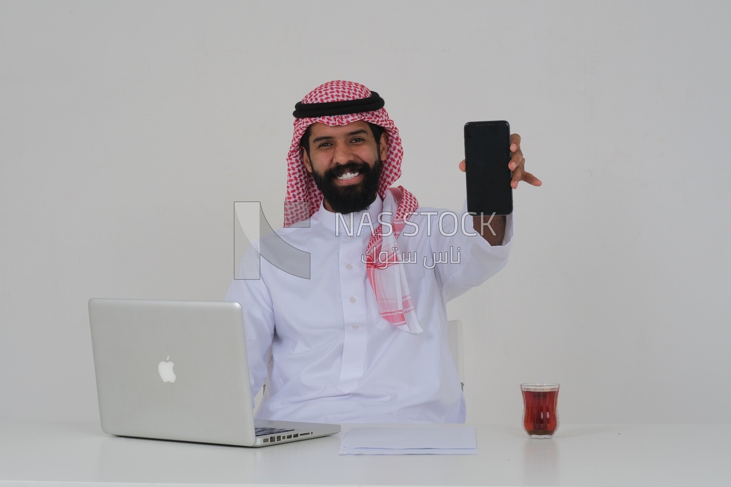 Saudi man sitting in front of a laptop, a cup of tea, using his mobile phone, business feasibility study, white background, Saudi model