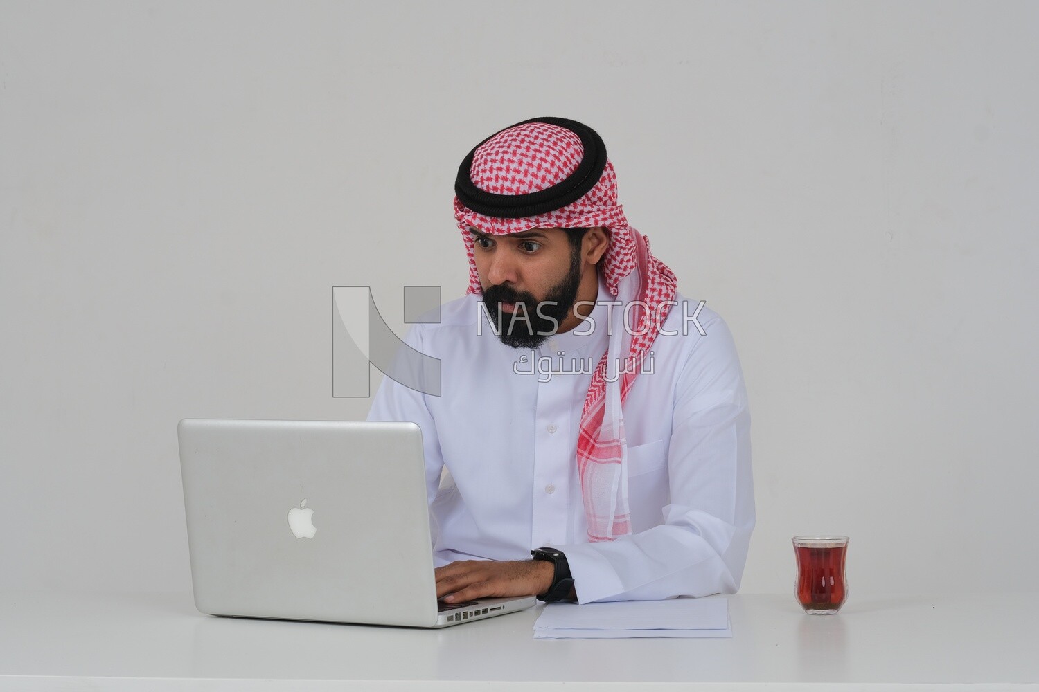 رجل سعودي يجلس ويعمل على جهاز كمبيوتر محمول ، فنجان شاي ، دراسة جدوى أعمال ، خلفية بيضاء ، موديل سعودي