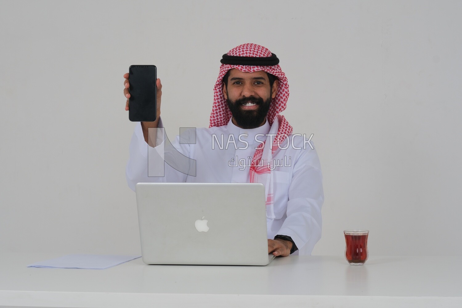 Saudi man sitting in front of a laptop, a cup of tea, using his mobile phone, business feasibility study, white background, Saudi model