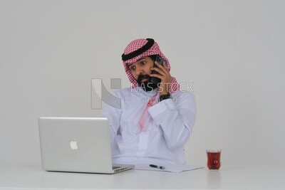 رجل سعودي يجلس ويعمل على جهاز كمبيوتر محمول ، يستخدم هاتفه المحمول ،  فنجان شاي ، دراسة جدوى أعمال ، خلفية بيضاء ، موديل سعودي